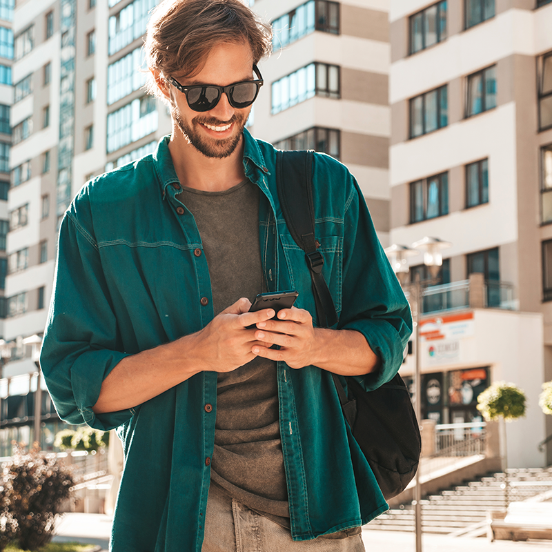 homem jovem sorrindo olhando para o celular. O homem está usando uma jaqueta preta e óculos